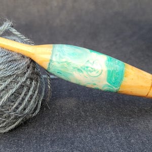 May include: A wooden crochet hook with a teal and white resin inlay. The hook is resting on a ball of gray yarn.