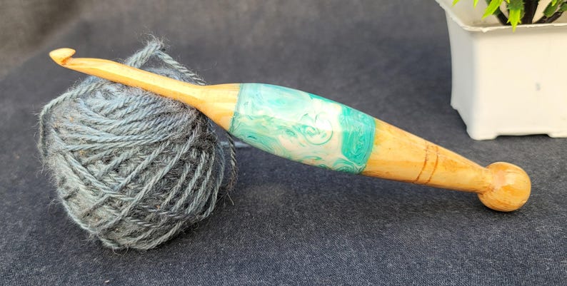 May include: A wooden crochet hook with a teal and white resin inlay. The hook is resting on a ball of gray yarn.