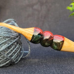 May include: A wooden crochet hook with a red, green, and black marbled design. The hook is resting on a ball of blue yarn.