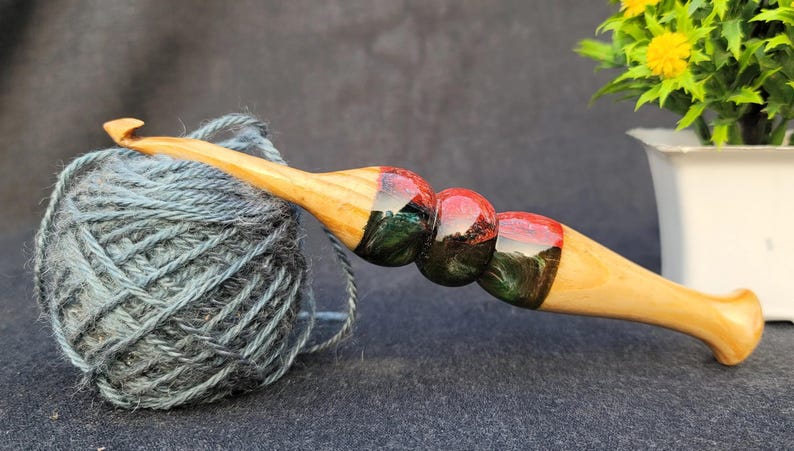 May include: A wooden crochet hook with a red, green, and black marbled design. The hook is resting on a ball of blue yarn.