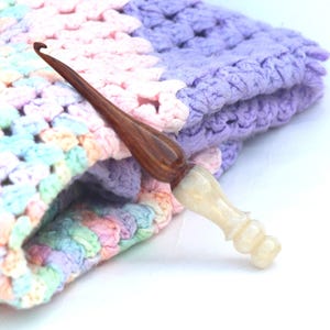 May include: A close-up of a crochet blanket in shades of pink, purple, and blue with a wooden and white crochet hook resting on top.