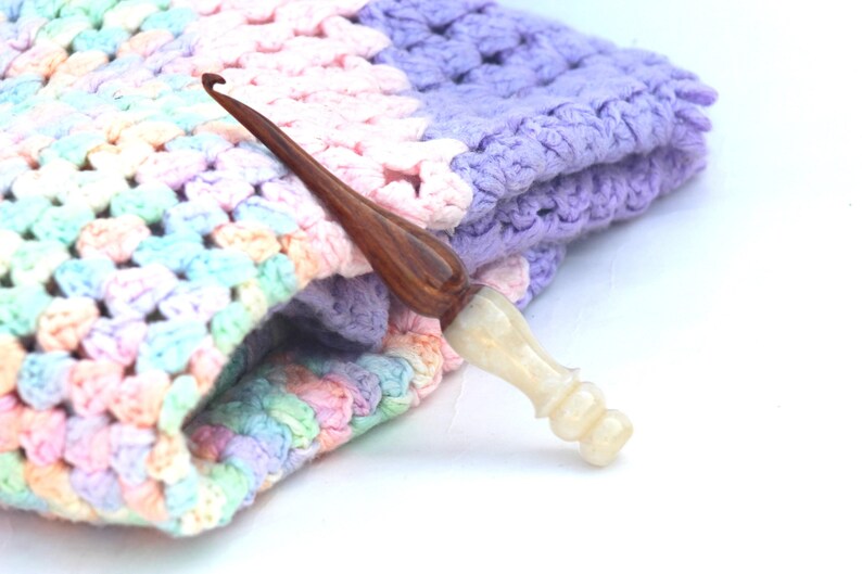 May include: A close-up of a crochet blanket in shades of pink, purple, and blue with a wooden and white crochet hook resting on top.
