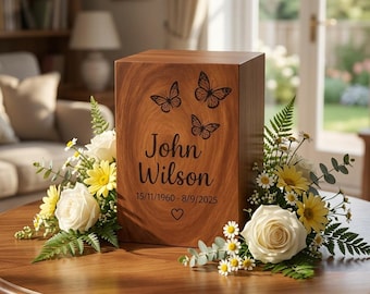Puede incluir: Una urna funeraria rectangular de madera con acabado de veta natural, grabada con tres mariposas, el nombre John Wilson, las fechas 15/11/1960-8/9/2025 y un pequeño corazón, rodeada de rosas blancas y margaritas amarillas.