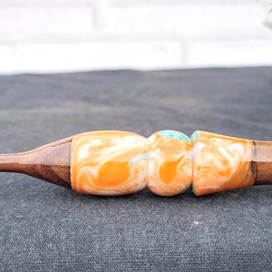 May include: A wooden crochet hook with a marbled orange and white handle. The handle has a teal swirl design.