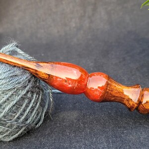 May include: A wooden crochet hook with a red and brown finish. The hook is resting on a ball of gray yarn.