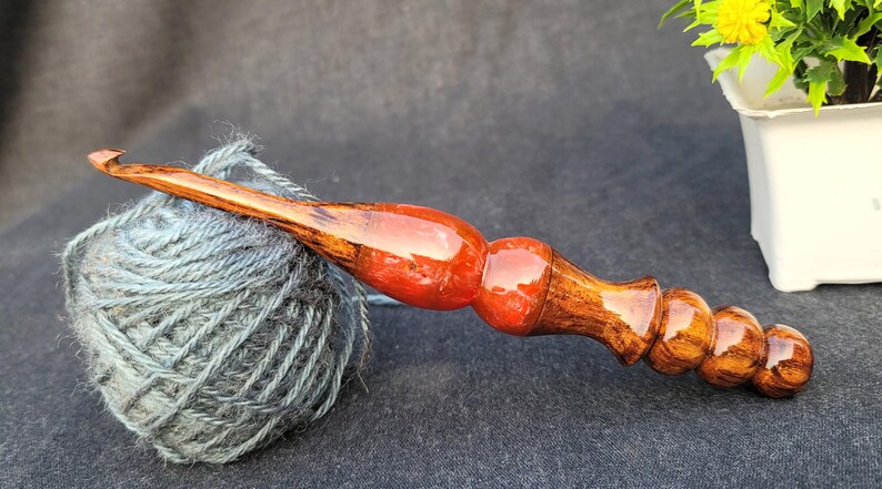 May include: A wooden crochet hook with a red and brown finish. The hook is resting on a ball of gray yarn.