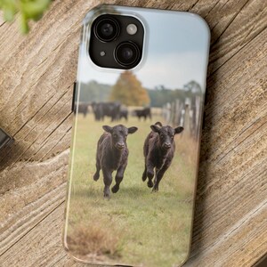 May include: A phone case featuring a pastoral scene with two black calves running across a grassy field. The background includes a wooden fence, trees, and a cloudy sky. The case is on a wooden surface.