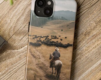 May include: A phone case featuring a landscape scene of a cowboy on horseback herding cattle. The case has a clear border and is resting on a wooden surface. A camera and greenery are also visible.