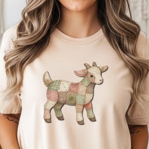 May include: A beige t-shirt with a patchwork goat illustration. The goat design incorporates pastel shades of pink, green, and beige. The t-shirt has a crew neck and short sleeves.