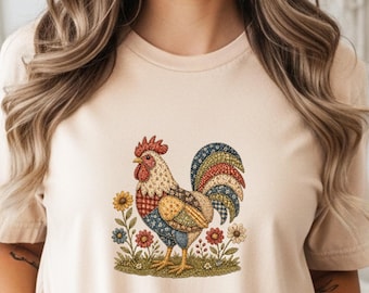 May include: A beige t-shirt featuring a colorful rooster design. The rooster is depicted with a red comb, a yellow and white body, and a blue and green tail. The design is surrounded by flowers and green grass.