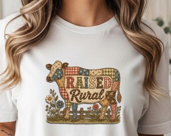 May include: White t-shirt featuring a colorful cow graphic with the text "RAISED RURAL". The cow design incorporates plaid and floral patterns. The shirt is made of a soft material and is suitable for casual wear.