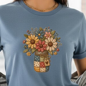 May include: A light blue t-shirt featuring a colorful floral arrangement in a patchwork mason jar design. The flowers include yellow, red, and blue blooms. The jar has a heart-shaped tag. The shirt has rolled-up sleeves.