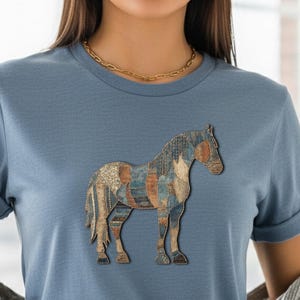 May include: A light blue t-shirt featuring a detailed horse graphic. The horse design incorporates various textures and colors, including shades of brown, blue, and gold. The shirt has short sleeves and a crew neck.