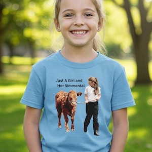 May include: A light blue t-shirt featuring the text "Just A Girl and Her Simmental" above an illustration of a girl with a cow. The girl is holding a leash attached to the cow. The background is a green grassy area.