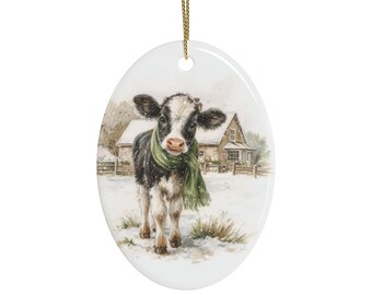 May include: An oval ceramic ornament featuring a black and white calf wearing a green scarf, standing in a snowy field with a house in the background. The ornament is white with a gold string attached at the top for hanging on a tree or display.