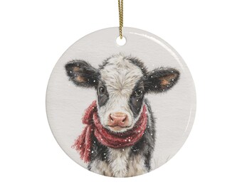 May include: A round ceramic ornament featuring a detailed illustration of a black and white calf wearing a textured red scarf. The calf is depicted against a light background with small white specks resembling falling snow. A gold string is attached.