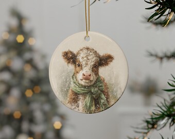 May include: A round ceramic ornament featuring a detailed illustration of a fluffy brown and white calf wearing a green scarf. The calf is depicted amidst light snowfall. The ornament hangs from a thin gold string against a blurred background.