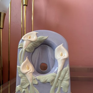 Könnte beinhalten: Ein dekoratives, bogenförmiges Keramikstück in Lavendel-, Creme- und Grüntönen. Es zeigt drei Calla-Lilienblüten mit detaillierten Blättern. Der Sockel hat einen gelben Rand. Der Hintergrund umfasst eine rosa Wand und goldene Lampen.