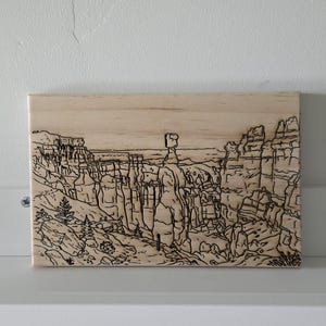 May include: A rectangular light-colored wooden plaque featuring a detailed black line art wood burning of a desert canyon landscape. The design depicts tall rock formations, including a prominent central hoodoo, with sparse vegetation.