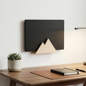 May include: A black rectangular tablet or display screen mounted on a wall, held by a light-colored wooden stand shaped like a mountain peak. The device features a smooth, matte black surface with a small circular button in the bottom left corner.