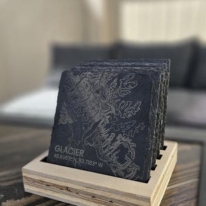 May include: A stack of square black slate coasters featuring an etched topographic map design. The front coaster displays the text GLACIER and the coordinates 48.6967 N, 113.7183 W. The coasters are held in a light-colored wooden holder with slots.