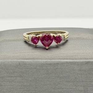 Puede incluir: Un anillo de oro con tres piedras de color rosa vibrante en forma de corazón dispuestas en fila. El corazón central es más grande que los dos laterales. Pequeñas piedras redondas y transparentes están incrustadas en el aro de oro.