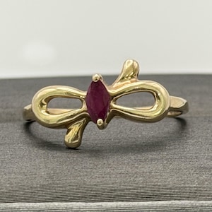 May include: A gold ring with a marquise-cut ruby gemstone at its centre. The ring has a distinctive bow-shaped design with swirling elements. The ring is set against a neutral background.