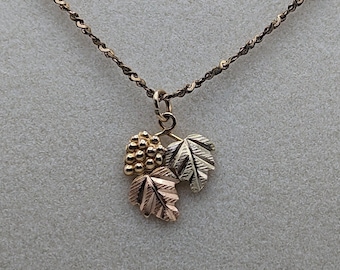 May include: A gold necklace with a pendant featuring three leaves and a cluster of grapes. The leaves are made of different shades of gold, and the grapes are made of yellow gold.