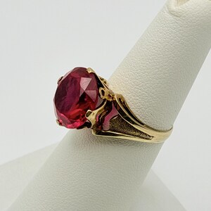 Op de afbeelding: Een gouden ring met een grote, ovaal geslepen, dieprode edelsteen in een klauwzetting. De ringband heeft een getextureerde, matte afwerking aan de zijkanten, die naar de centrale steen leidt. Het metaal lijkt gepolijst geelgoud te zijn.