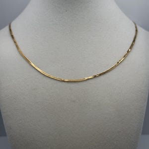 Puede incluir: Un collar de cadena de espiga en tono dorado con un diseño plano y flexible que descansa en una curva suave. La superficie metálica tiene un acabado pulido y reflectante que capta la luz, creando un brillo metálico uniforme en los eslabones.