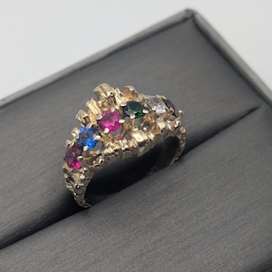 Puede incluir: Un anillo de oro con un diseño de corona que presenta múltiples piedras preciosas de colores, incluyendo rojo, rosa, azul, verde y blanco.