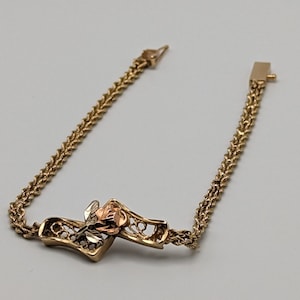 Puede incluir: Un brazalete de cadena de oro con un dije de rosa. La rosa está hecha de tres colores de oro diferentes: amarillo, blanco y rosa.