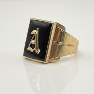 Könnte beinhalten: Ein goldfarbener Ring mit einer schwarzen, rechteckigen Fassung. Die Fassung zeigt einen goldenen Buchstaben "A". Der Ring hat ein klassisches, kühnes Design. Der Ring ist wahrscheinlich aus Metall und wird auf einer reflektierenden Oberfläche präsentiert.