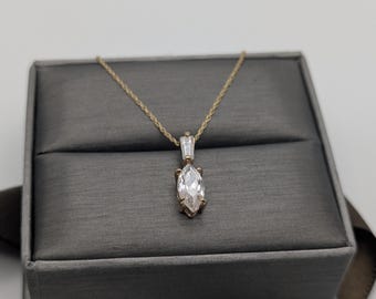 May include: A gold necklace featuring a marquise-cut pendant with a clear gemstone. The pendant hangs from a fine gold chain. The necklace is presented in a grey jewellery box.