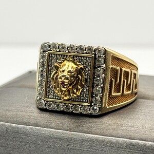 May include: A gold-toned ring featuring a square face with a central sculpted lion head in relief. The lion is surrounded by a border of small clear stones set in a textured silver-toned square. The sides of the ring display a geometric Greek key pattern.