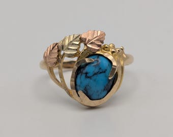 May include: A gold ring with a large, oval turquoise stone. The ring has a decorative design with gold leaves and accents. The turquoise stone is a vibrant blue colour, and the ring is set against a neutral background.