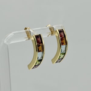 May include: A pair of curved gold hoop earrings featuring a channel setting with a row of multi-colored square-cut gemstones. The stones display a gradient of colors including deep red, amber, light blue, green, and purple set in polished gold.