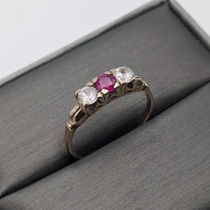 May include: A gold ring with three stones. The center stone is a red ruby, and the two stones on either side are clear diamonds.