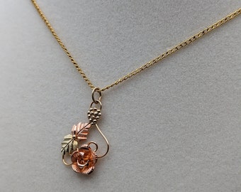 May include: A gold chain necklace with a pendant featuring a rose with green and gold leaves. The rose is made of three different shades of gold.