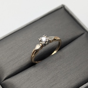 May include: A gold ring with a single round diamond set in a prong setting. The ring has a delicate design with two small heart-shaped accents on either side of the diamond.