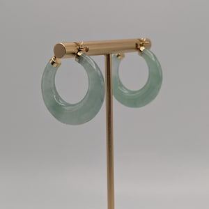 Op de afbeelding: Een paar jade oorringen. De oorbellen zijn lichtgroen van kleur en hebben een glad, gepolijst oppervlak. Ze worden gepresenteerd op een goudkleurige oorringstandaard tegen een neutrale achtergrond.