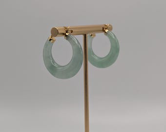 May include: A pair of jade hoop earrings. The earrings are a light green color and have a smooth, polished surface. They are displayed on a gold-colored earring stand against a neutral background.