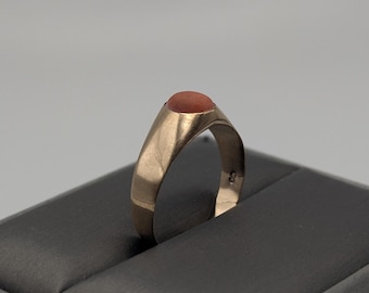 May include: A gold-colored ring with a rounded, reddish-orange stone set in a smooth, polished band. The ring is displayed on a dark gray surface, showcasing its simple yet elegant design. The ring's band has a slight curve.