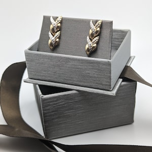 Puede incluir: Un par de pendientes de oro y plata en una caja de regalo gris con una cinta marrón. Los pendientes tienen forma de espiga de trigo con pequeños diamantes.