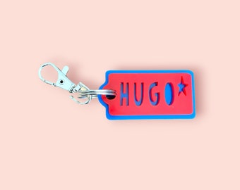 May include: A red and blue keychain with the name "HUGO" cut out. A blue star is on the right side of the keychain. The keychain has a silver clasp.