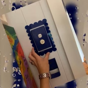 Puede incluir: Una tapa de interruptor de luz azul con un borde festoneado. La tapa está montada en una pared blanca.