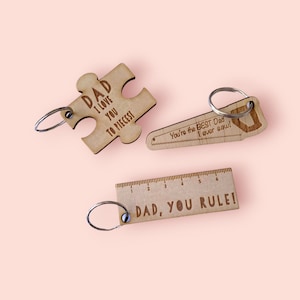 May include: Three wooden keychains with different designs. The first keychain is a jigsaw piece with the text "DAD I LOVE YOU TO PIECES". The second keychain is a saw with the text "You're the BEST Dad I ever saw!". The third keychain is a ruler with the text "DAD, YOU RULE!"