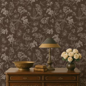 May include: A dark brown floral wallpaper with white sketched botanical designs. A wooden dresser with a lamp, books, a decorative bowl, and a vase of white roses sits in front of the wallpaper.
