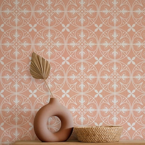 May include: A peach-colored wall with a white, ornate, repeating pattern. A brown vase with a circular opening holds a dried palm leaf. A woven basket sits on a wooden surface, complementing the warm tones of the background.