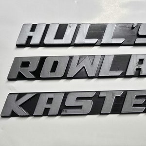 May include: Three metallic nameplates with the names "HULL'S", "ROWLAND", and "KASTEN" in a sans-serif font. The letters are silver against a black background. The nameplates are arranged horizontally on a white surface.